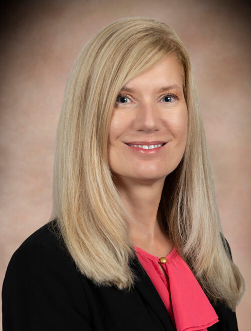 Smiling blonde woman in a black blazer and pink blouse, head-and-shoulders portrait.